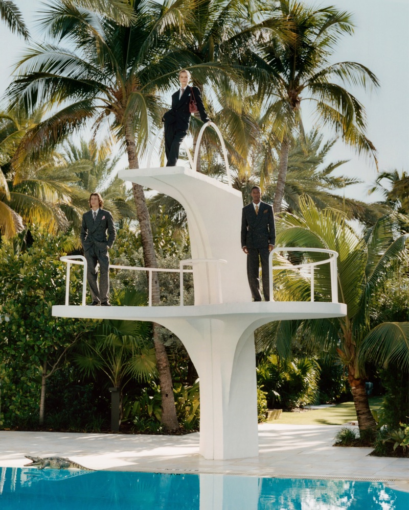 Three models in dark suits posed on a white multi-level diving board next to a pool surrounded by palm trees.