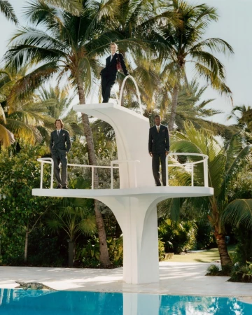 Three models in dark suits posed on a white multi-level diving board next to a pool surrounded by palm trees.