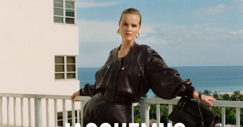 Model Eva Herzigova leaning on a railing against an ocean view, wearing a dark leather jacket and holding a black handbag.