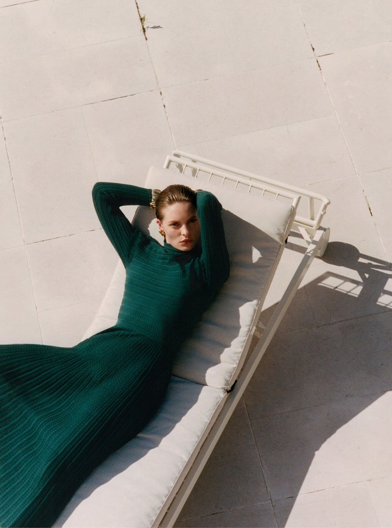 Model wearing a dark green ribbed dress reclining on a white lounge chair outdoors on tile paving.