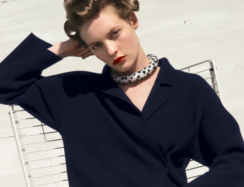 Model with styled hair wearing a dark navy collared top and beaded necklace seated outdoors against a bright white background