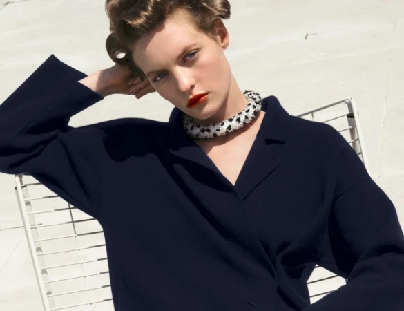 Model with styled hair wearing a dark navy collared top and beaded necklace seated outdoors against a bright white background