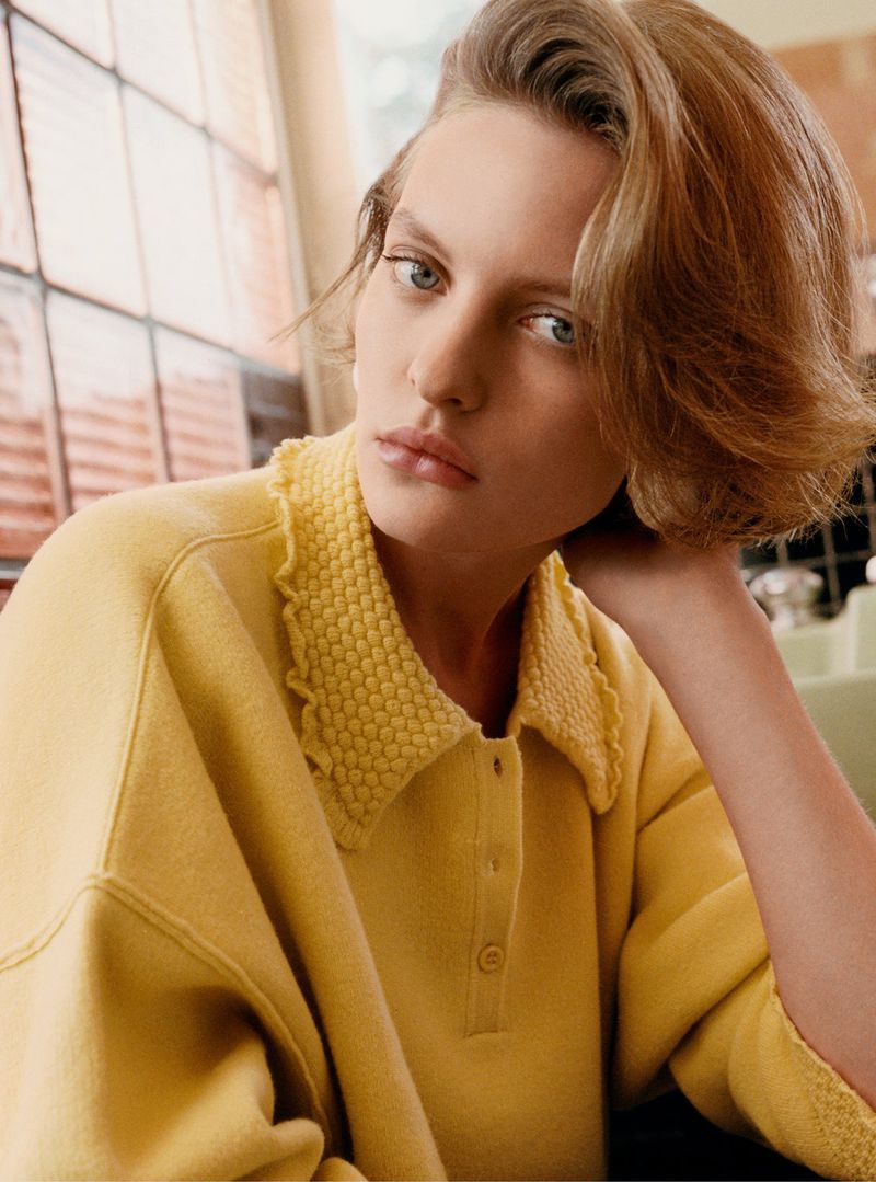 Model wearing a yellow knit collared polo top with textured collar detail posing near a window.