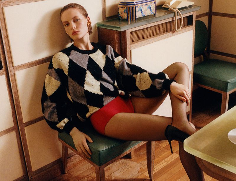 Model seated on a green chair wearing an argyle sweater and red shorts inside an interior space.