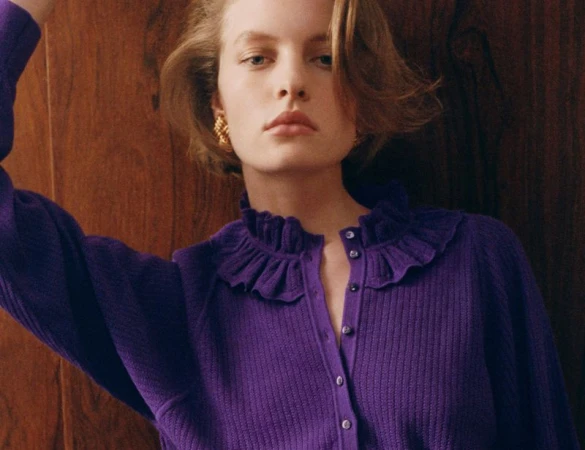 Model in a purple ribbed cardigan with a ruffled collar posing against a dark wood paneled wall.