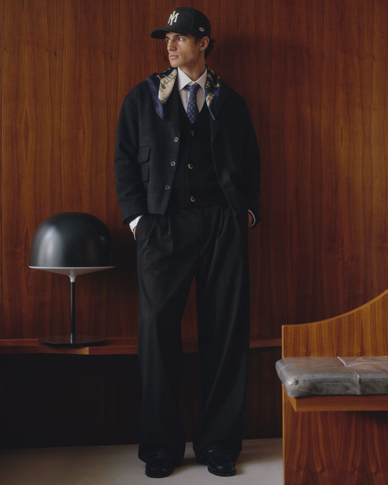 Man wearing a dark baseball cap, black jacket, trousers, and a patterned scarf standing against a wood paneled wall.