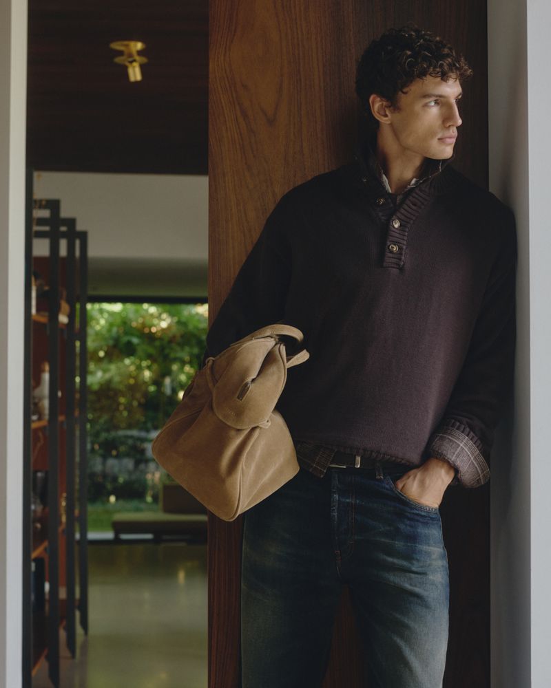 Man with curly brown hair holding a tan suede bag while leaning against a wooden door frame in a domestic interior