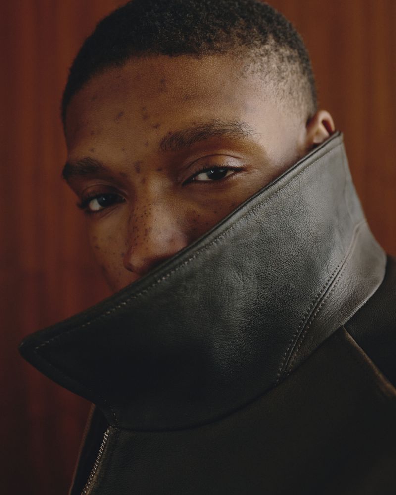 Close-up portrait of a young Black man wearing a high-collared dark leather jacket against a wood-paneled wall