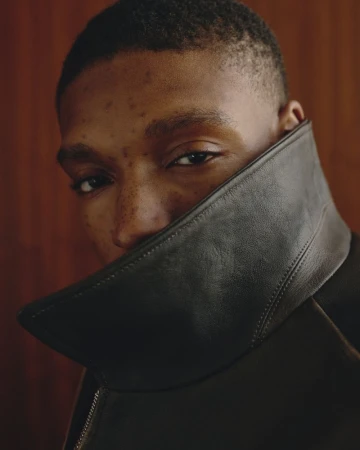 Close-up portrait of a young Black man wearing a high-collared dark leather jacket against a wood-paneled wall