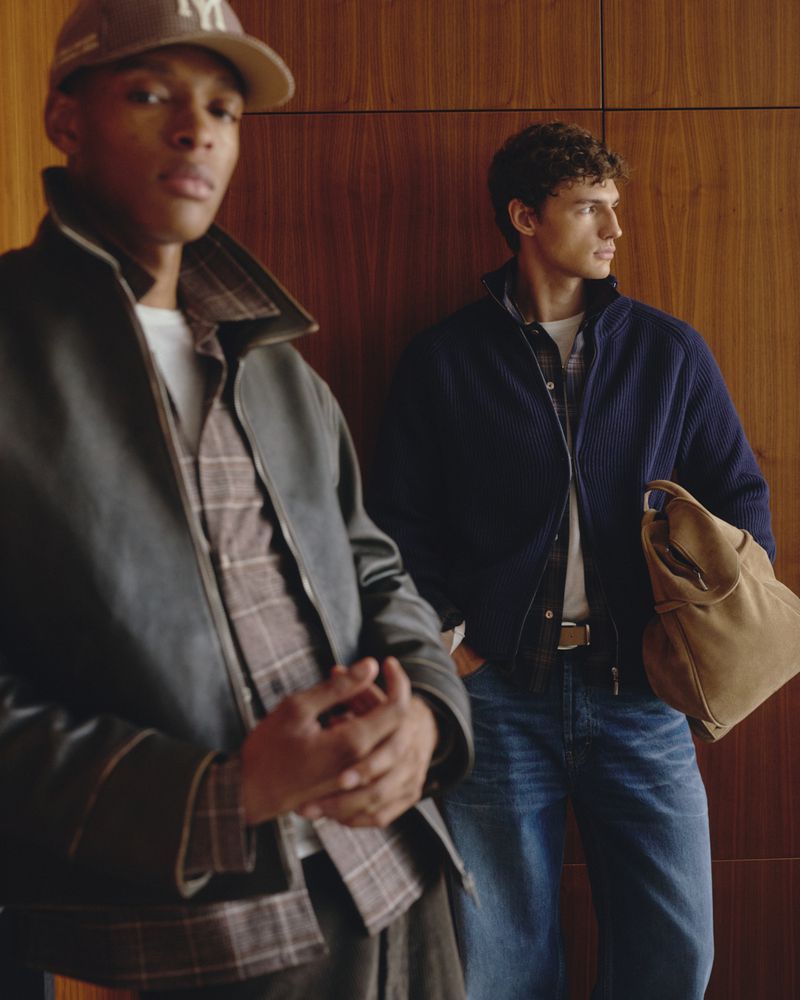 Two male models posing indoors with wood paneling walls, wearing layered jackets, blue jeans, and carrying a tan bag.