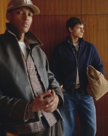 Two male models posing indoors with wood paneling walls, wearing layered jackets, blue jeans, and carrying a tan bag.