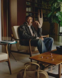 Man seated in a mid-century armchair wearing a dark leather jacket, holding an ornate wooden object inside a library.