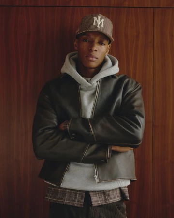 A man wearing a dark leather jacket, grey hoodie, and patterned baseball cap standing against wood panelling.