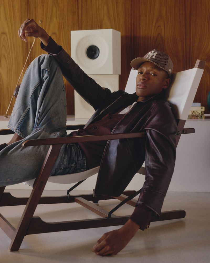 A Black man seated in a white sling lounge chair wearing a brown leather jacket, baseball cap, and wide-leg jeans inside a wood-paneled room
