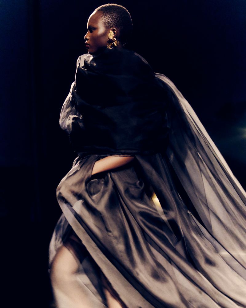 Model wearing a dark draped gown and gold earring against a dark background, photographed by Lucie Rox for Saint Laurent SS27.