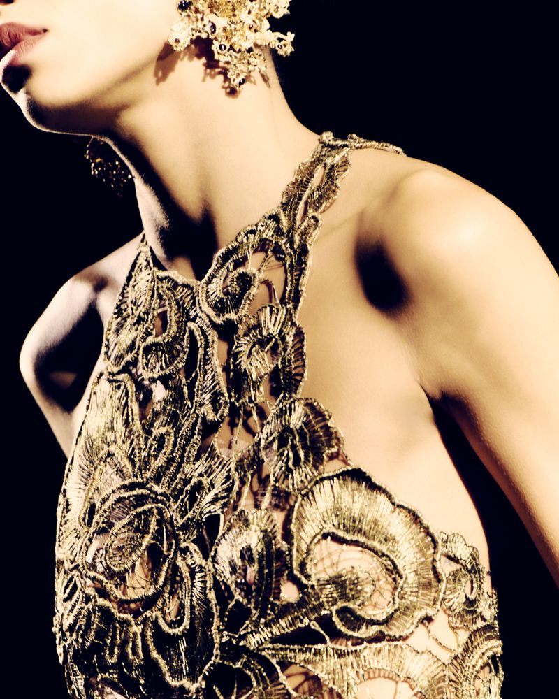 Close-up portrait of a model wearing an ornate gold embroidered halter top and intricate earrings against a black background.