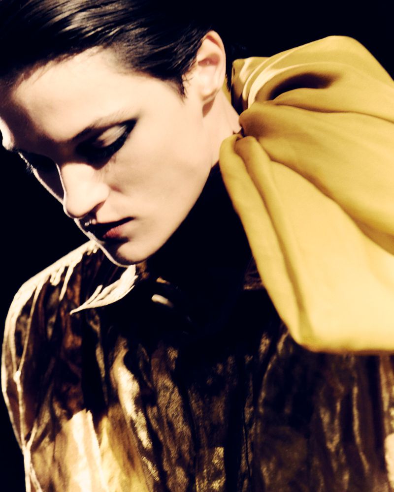 Close-up portrait of a model with dark slicked-back hair wearing a glossy metallic top against a dark background