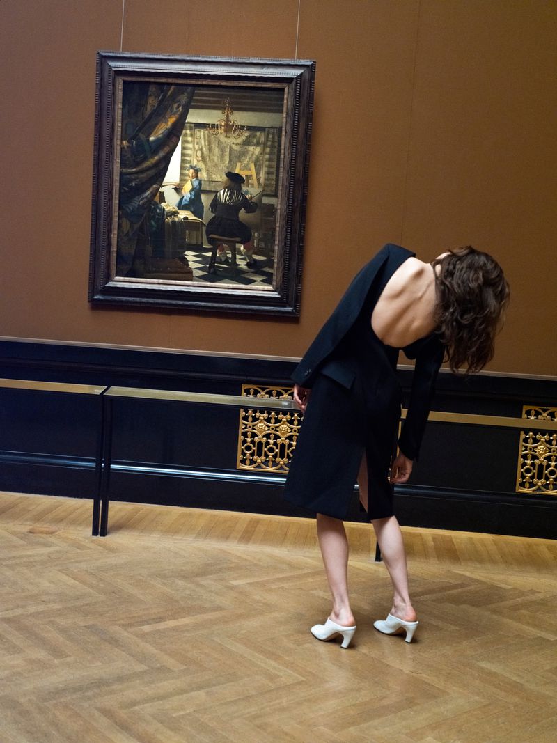 Model Anna de Rijk wearing a black backless dress and white mules posing in a gallery with a painting.