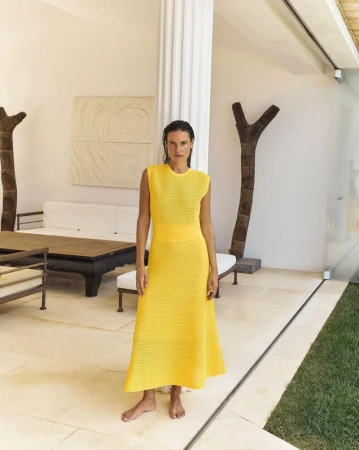 Model wearing a bright yellow sleeveless knit dress standing barefoot inside a modern light-filled interior