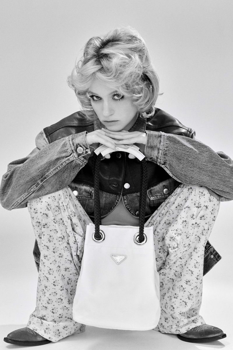 Black and white portrait of a woman in denim jacket, floral pants, and western boots holding a Prada handbag in a photo studio.