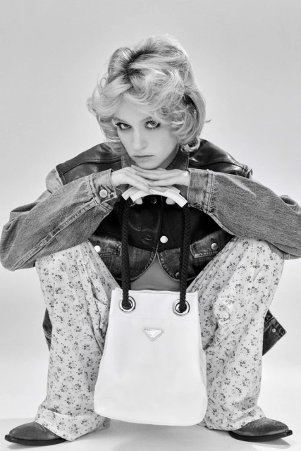Black and white portrait of a woman in denim jacket, floral pants, and western boots holding a Prada handbag in a photo studio.