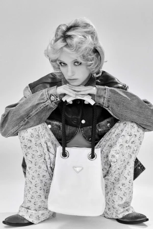 Black and white portrait of a woman in denim jacket, floral pants, and western boots holding a Prada handbag in a photo studio.