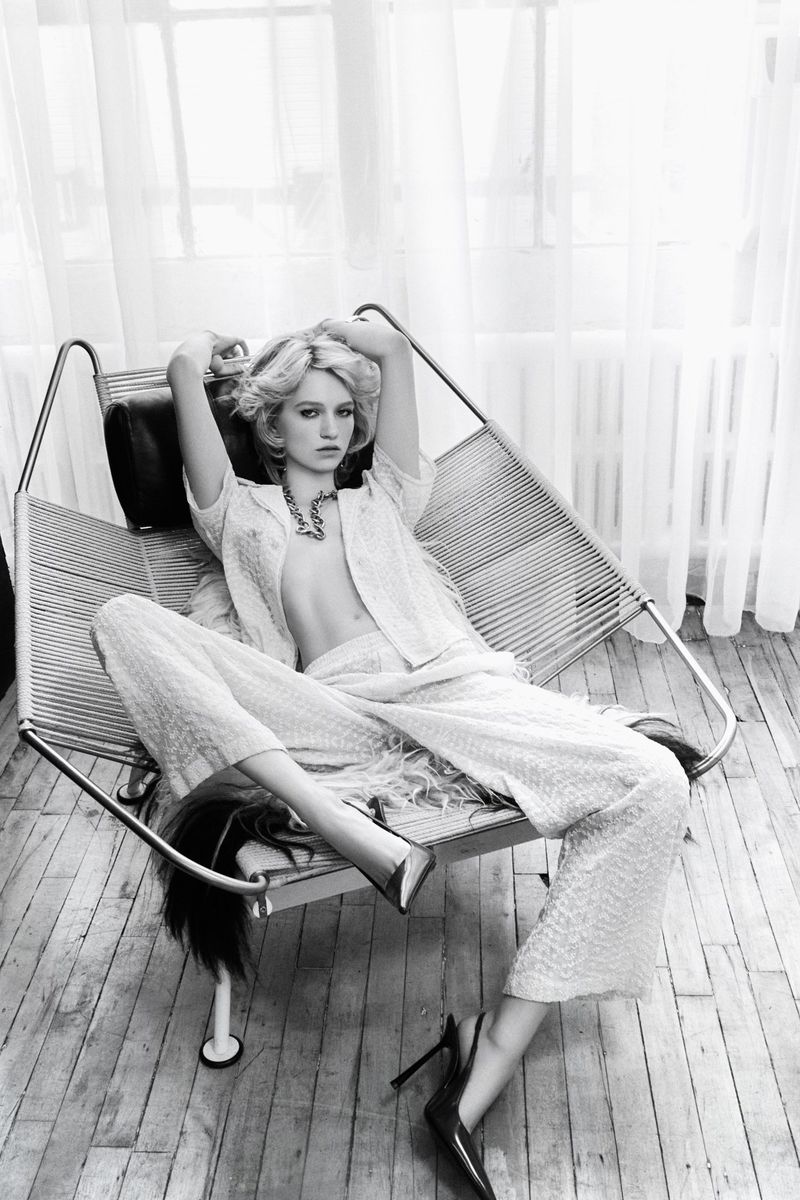 A blonde woman with an unbuttoned shirt and pants poses in a metal lounge chair in a studio with neutral colors and soft light.