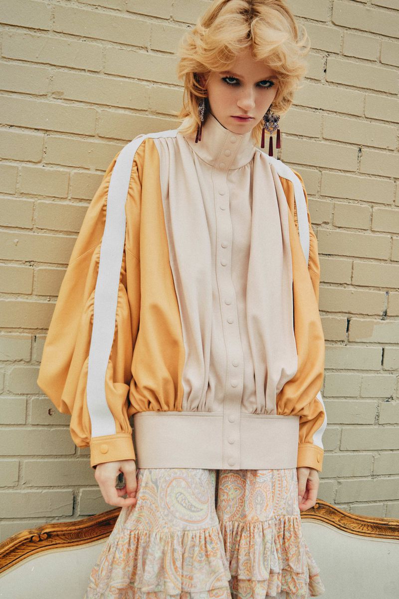Blonde woman in high-neck blouse and paisley pants stands against brick wall, wearing statement earrings in fashion editorial shot.
