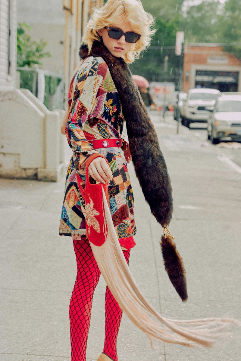 Fashionable woman in a colorful patchwork coat, fishnet stockings, and an unusual red handbag walks along a city street on a rainy day.