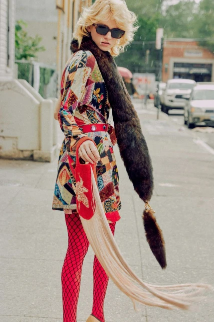 Fashionable woman in a colorful patchwork coat, fishnet stockings, and an unusual red handbag walks along a city street on a rainy day.