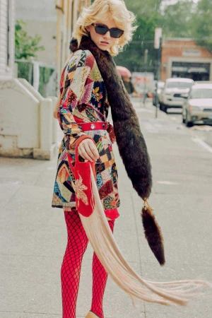 Fashionable woman in a colorful patchwork coat, fishnet stockings, and an unusual red handbag walks along a city street on a rainy day.
