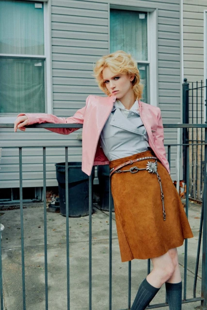 Adult female fashion model poses in brown suede skirt, pink jacket, gray top and knee socks by gray clapboard house with fence.