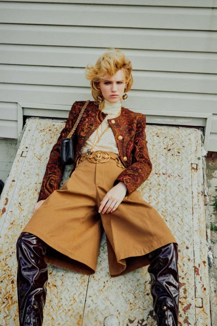 Fashion shot of a woman in wide-leg trousers and paisley jacket posing against a metal door, accessorized with jewelry and a purse.
