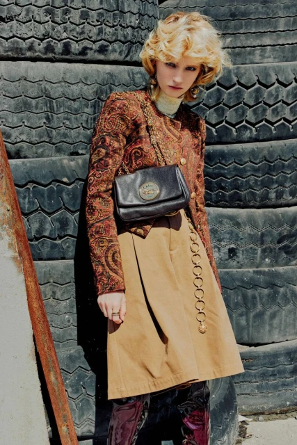 Woman in patterned jacket, tan skirt, and boots poses against a wall of stacked tires, showcasing fashion and style in an outdoor setting.