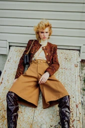 Model with blonde hair wearing patterned jacket and wide-legged tan trousers seated against a pale building exterior