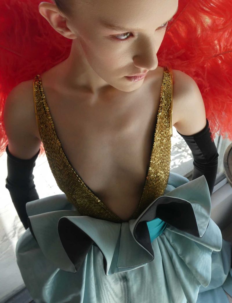High-angle fashion editorial portrait of a model in a gold sequin top, light blue skirt, and large red feathers.