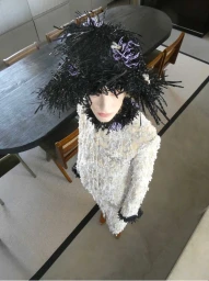 High-angle fashion editorial shot of a model wearing a textured white coat and a large black fringe hat indoors