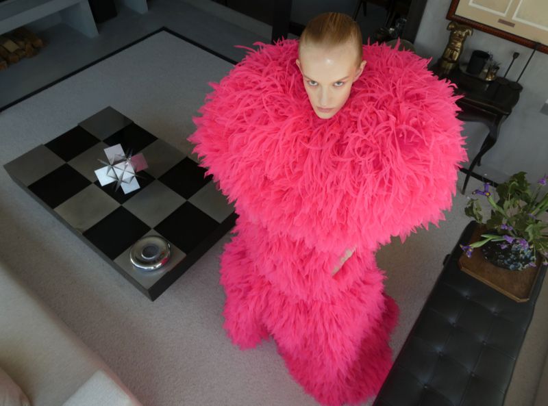 Model wearing a voluminous bright pink feathered garment looking up in an interior room viewed from above.