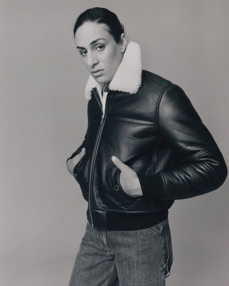 Portrait of Imane Khelif wearing a black leather jacket with a white shearling collar and denim jeans against a plain neutral background.