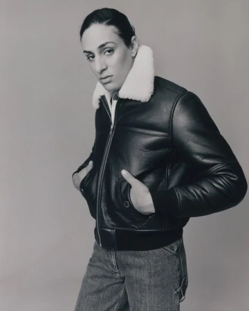 Portrait of Imane Khelif wearing a black leather jacket with a white shearling collar and denim jeans against a plain neutral background.