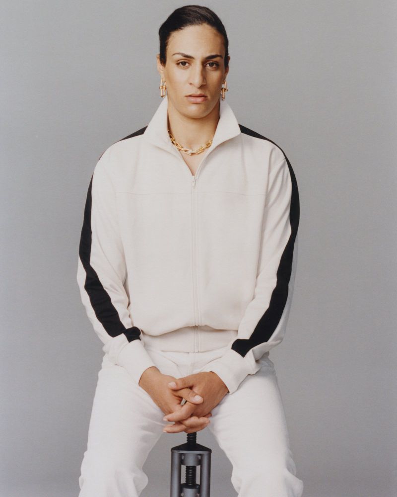 Portrait of Imane Khelif wearing a white tracksuit with black stripes against a grey backdrop for Mastermind Magazine