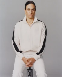 Portrait of Imane Khelif wearing a white tracksuit with black stripes against a grey backdrop for Mastermind Magazine