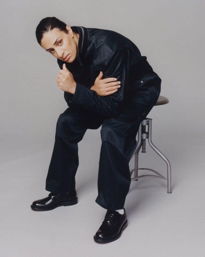 Imane Khelif seated on a metal stool wearing a dark utility jumpsuit and black leather shoes against a grey background