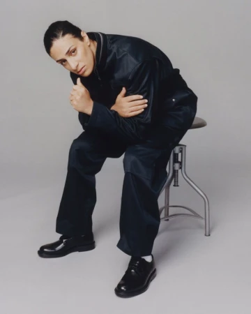 Imane Khelif seated on a metal stool wearing a dark utility jumpsuit and black leather shoes against a grey background