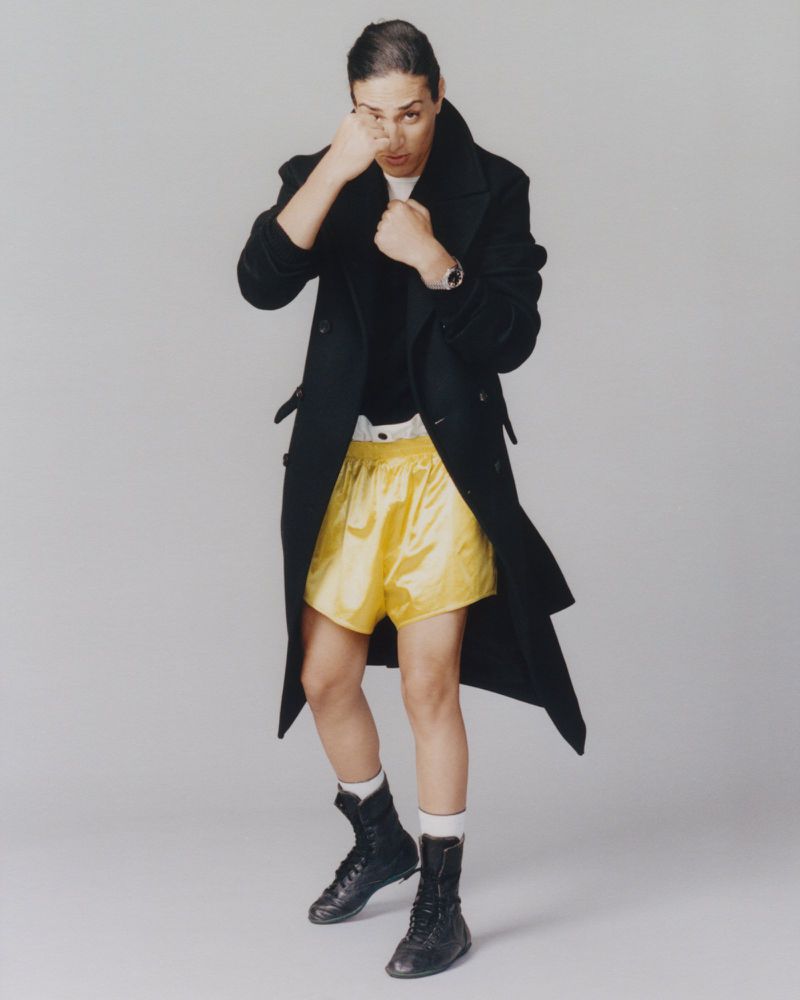 Imane Khelif in a black coat and yellow boxing shorts holding up her fists against a grey background