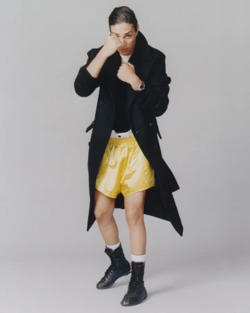 Imane Khelif in a black coat and yellow boxing shorts holding up her fists against a grey background