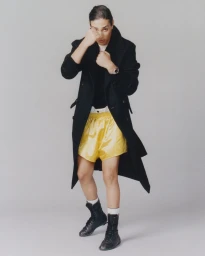 Imane Khelif in a black coat and yellow boxing shorts holding up her fists against a grey background