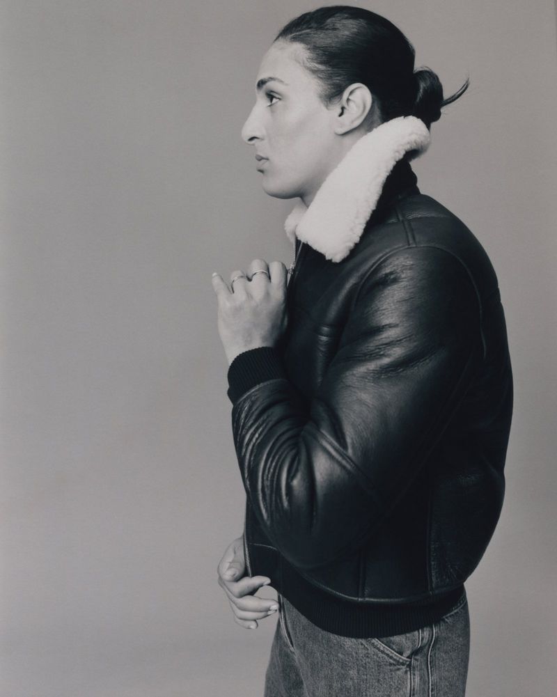 Portrait profile of Imane Khelif wearing a black leather jacket with white shearling collar against a neutral studio background