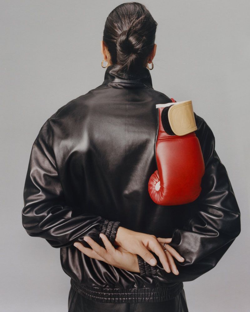Rear view of a person wearing a black leather jacket with a red boxing glove tucked over the shoulder against a grey studio background