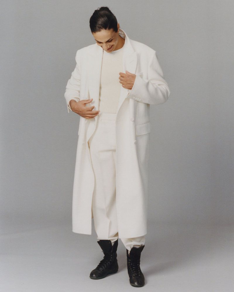 Imane Khelif wearing a white long coat and black boots against a neutral grey studio background for Mastermind Magazine.